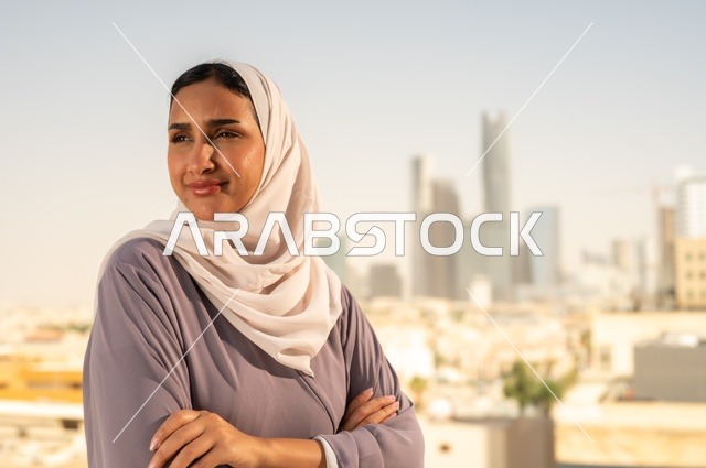الاناقة والاهتمام بالمظهر الخارجي، ،صورة  مقربة لشابة عربية خليجية سعودية ترتدي العباءة تقف بايدي متقاطعة ، النظر الى شيء ما بايماءات السعادة والسرور،الخروج في الهواء الطلق