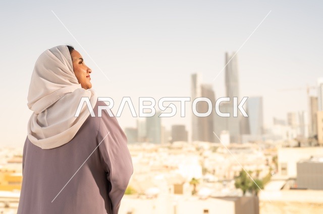 الافتخار بالانتماء للمملكه ، مفهوم الاناقة والاهتمام بالمظهر الخارجي،صورة من الخلف لشابة عربية خليجية سعودية ترتدي العباءة، النظر الى شيء ما بايماءات السعادة والسرور،الخروج في الهواء الطلق