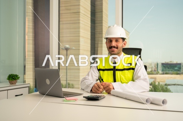 الإمساك بمخططات الإعمار، صورة مقربة لمهندس عربي خليجي سعودي مبتسم يرتدي الثوب التقليدي والسترة والخوذة الخاصة بالعمل يجلس على المكتب ويمسك بيده المخطط الهندسي بجانبه الحاسب المحمول، متابعه وتدقيق سير المشروع, مفهوم الهندسة والإنشاءات
