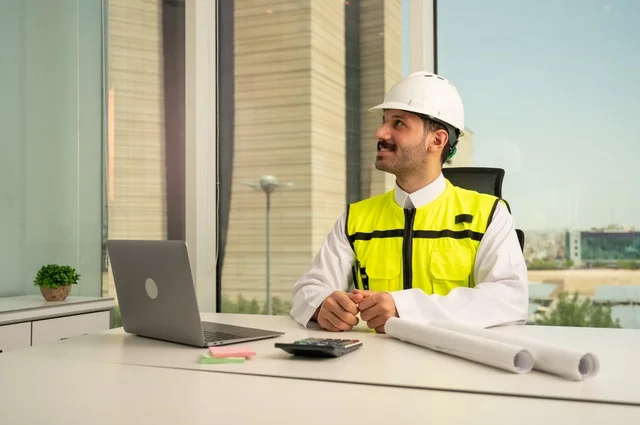  لإكمال رسم مخطوطات البناء دمج التكنولوجيا مع الهندسة ،تدقيق ومراجعة الخطط الهندسية, مفهوم الهندسة والإنشاءات، مهندس عربي خليجي سعودي يرتدي الثوب التقليدي والسترة والخوذة الخاصة بالعمل يجلس على المكتب ويعمل على الحاسوب المحمول 