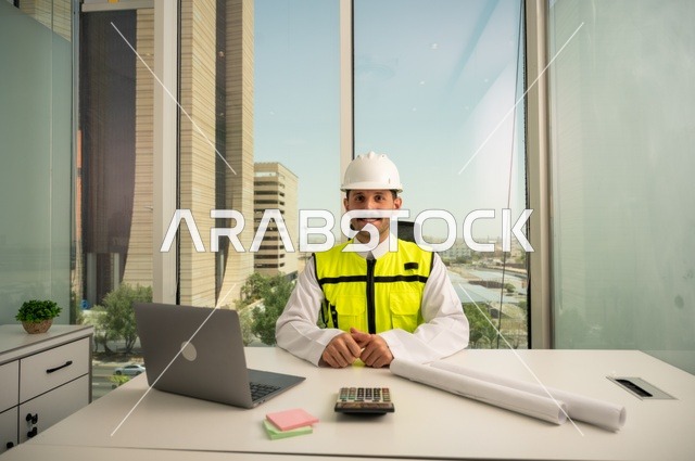   نظرة فخر واعتزاز للكاميرا، تدقيق ومراجعة الخطط الهندسية, دمج التكنولوجيا مع الهندسة ،مفهوم الهندسة والإنشاءات، مهندس عربي خليجي سعودي مبتسم يرتدي الثوب التقليدي والسترة الخاصة بالعمل يجلس على المكتب بجانبه حاسوب محمول ومخططات هندسية