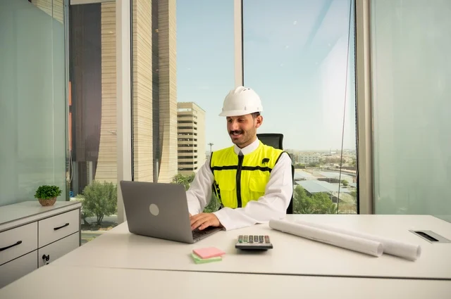 Integrating technology with engineering to complete construction drawings, review and audit engineering plans, engineering and construction concept, Saudi Arabian Gulf engineer wearing traditional thobe, jacket and work helmet sitting at desk working on laptop