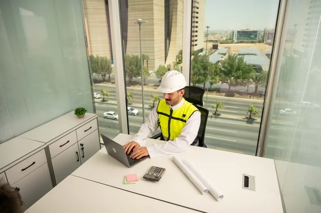  دمج التكنولوجيا مع الهندسة لإكمال رسم مخطوطات البناء،تدقيق ومراجعة الخطط الهندسية, مفهوم الهندسة والإنشاءات، مهندس عربي خليجي سعودي يرتدي الثوب التقليدي والسترة والخوذة الخاصة بالعمل يجلس على المكتب ويعمل على الحاسوب المحمول 