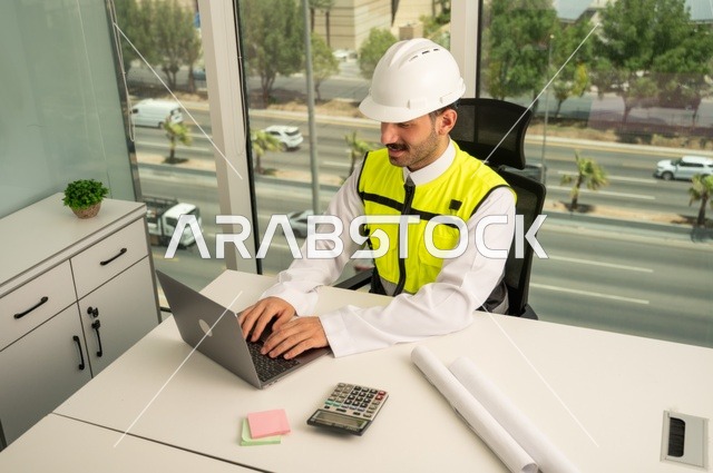  لإكمال رسم مخطوطات البناء دمج التكنولوجيا مع الهندسة ،تدقيق ومراجعة الخطط الهندسية, مفهوم الهندسة والإنشاءات، مهندس عربي خليجي سعودي يرتدي الثوب التقليدي والسترة والخوذة الخاصة بالعمل يجلس على المكتب ويعمل على الحاسوب المحمول 