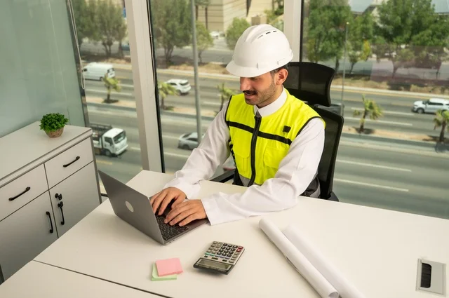  لإكمال رسم مخطوطات البناء دمج التكنولوجيا مع الهندسة ،تدقيق ومراجعة الخطط الهندسية, مفهوم الهندسة والإنشاءات، مهندس عربي خليجي سعودي يرتدي الثوب التقليدي والسترة والخوذة الخاصة بالعمل يجلس على المكتب ويعمل على الحاسوب المحمول 