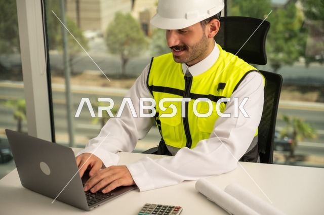   مهارة وسرعة الأيدي في الكتابة، تدقيق ومراجعة الخطط الهندسية, لإكمال رسم مخطوطات البناء دمج التكنولوجيا مع الهندسة ،مفهوم الهندسة والإنشاءات، مهندس عربي خليجي سعودي يرتدي الثوب التقليدي والسترة الخاصة بالعمل يجلس على المكتب ويعمل على الحاسوب المحمول 