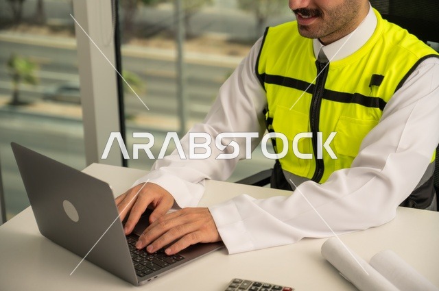   مهارة وسرعة الأيدي في الكتابة، تدقيق ومراجعة الخطط الهندسية, لإكمال رسم مخطوطات البناء دمج التكنولوجيا مع الهندسة ،مفهوم الهندسة والإنشاءات، مهندس عربي خليجي سعودي يرتدي الثوب التقليدي والسترة الخاصة بالعمل يجلس على المكتب ويعمل على الحاسوب المحمول 