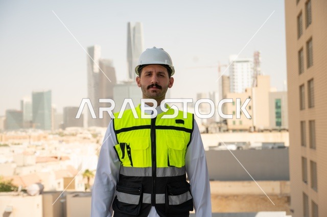 العمل في المجال الهندسي، الوقوف بفخر واعتزاز والنظر إلى الكاميرا, صورة مقربة لمهندس عربي خليجي سعودي مبتسم يرتدي سترة العمل وخوذة الحماية البيضاء, ، الاهتمام بالتطور العمراني بسواعد المهندسين السعوديين، خلفية الأبراج وناطحات السحاب في مدينة الرياض