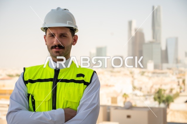 الاهتمام بالتطور العمراني بسواعد المهندسين السعوديين، الوقوف بفخر واعتزاز والنظر إلى الكاميرا, مهندس عربي خليجي سعودي مبتسم يرتدي سترة العمل وخوذة الحماية البيضاء,ايماءات أيدي متقاطعة, خلفية الأبراج وناطحات السحاب في مدينة الرياض