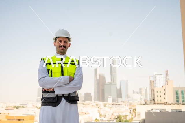 Taking care of urban development with the hands of Saudi engineers, standing proudly and looking at the camera, a smiling Saudi Arabian Gulf engineer wearing a work jacket and a white protective helmet, crossed hand gestures, background of towers and skyscrapers in the city of Riyadh