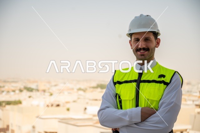  الوقوف بفخر واعتزاز والنظر إلى الكاميرا, التطور العمراني بسواعد المهندسين السعوديين، صورة مقربة لمهندس عربي خليجي سعودي مبتسم يرتدي سترة العمل وخوذة الحماية البيضاء,ايماءات أيدي متقاطعة, خلفية الأبراج وناطحات السحاب في مدينة الرياض