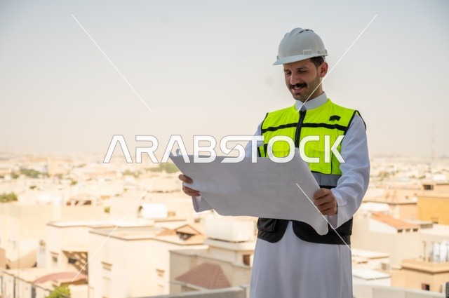 مفهوم وادرة المشاريع وتدقيقها، الأشراف على العمل لمتابعة سير المشاريع وتنفيذها، صورة جانبية لمهندس عربي خليجي سعودي يرتدي الثوب التقليدي والسترة الخاصة وخوذة الحماية يحمل مخطط هندسي، خلفية أبراج وناطحات سحاب