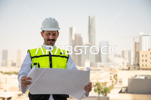 التطور والنمو العمراني على ايدي مهندسين الوطن، مفهوم الأشراف والتدقيق على العمل لمتابعة سير المشاريع وتنفيذها، صورة جانبية لمهندس عربي خليجي سعودي يرتدي الثوب التقليدي والسترة الخاصة وخوذة الحماية يحمل ويقرأ مخطط هندسي، خلفية أبراج وناطحات سحاب