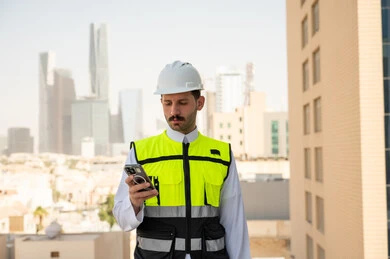 تطور ونمو القطاع الهندسي في المملكة، الإعمار بسواعد أبناء الوطن، مهندس عربي خليجي سعودي يرتدي الثوب التقليدي والسترة الخاصة وخوذة الحماية يقف ويمسك الهاتف المحمول يتابع العمل حسب استراتيجية سير العمل،مفهوم الهندسة والانشاءات المعمارية، خلفية أبراج وناطحات سحاب