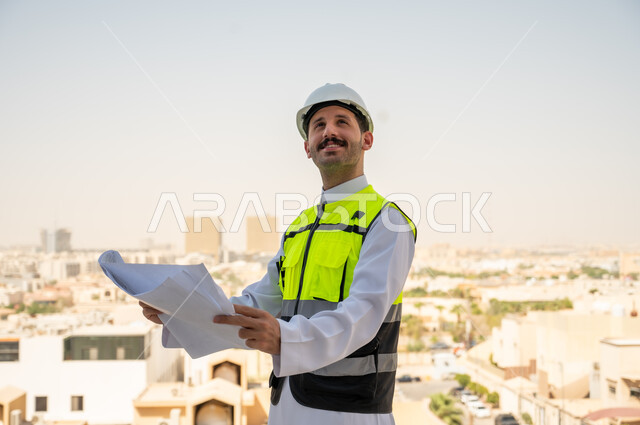  انجاز المهام اليومية لمتابعة سير المشاريع وتنفيذها، مفهوم أدارة المشاريع الهندسية، صورة جانبية لمهندس عربي خليجي سعودي يرتدي الثوب التقليدي والسترة الخاصة وخوذة الحماية يحمل ويقرأ مخطط هندسي، خلفية أبراج وناطحات سحاب في الرياض