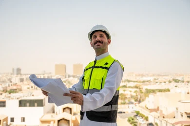  انجاز المهام اليومية لمتابعة سير المشاريع وتنفيذها، مفهوم أدارة المشاريع الهندسية، صورة جانبية لمهندس عربي خليجي سعودي يرتدي الثوب التقليدي والسترة الخاصة وخوذة الحماية يحمل ويقرأ مخطط هندسي، خلفية أبراج وناطحات سحاب في الرياض