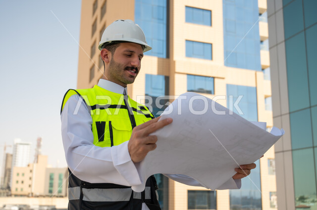 Supervising and auditing work to follow up on the progress of projects and their implementation, the concept of engineering project management, a side view of a Saudi Arabian Gulf engineer wearing traditional clothing, a special jacket and a protective helmet, holding and reading an engineering plan with gestures of thinking and planning, a background of a tower and skyscrapers