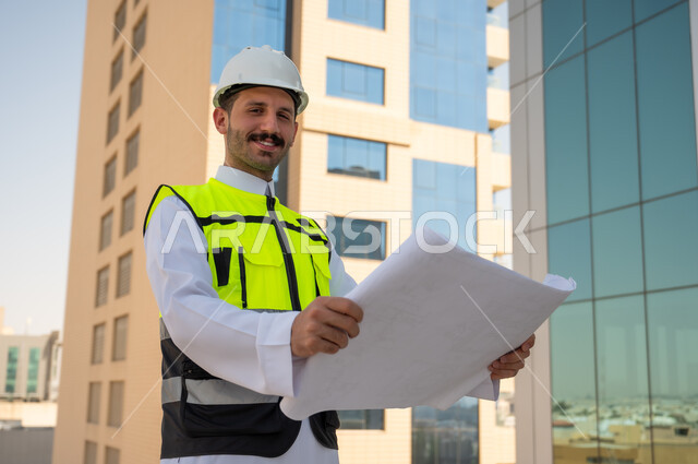 التطور والنمو العمراني على ايدي مهندسين الوطن، مفهوم الأشراف والتدقيق على العمل لمتابعة سير المشاريع وتنفيذها، صورة جانبية لمهندس عربي خليجي سعودي يرتدي الثوب التقليدي والسترة الخاصة وخوذة الحماية يحمل ويقرأ مخطط هندسي، خلفية أبراج وناطحات سحاب