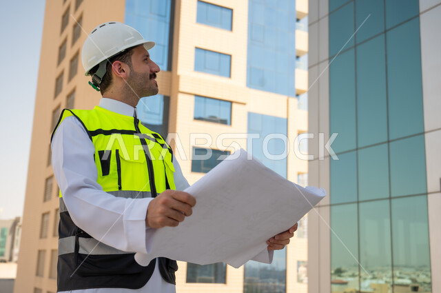 مفهوم وادرة المشاريع وتدقيقها، الأشراف على العمل لمتابعة سير المشاريع وتنفيذها، صورة جانبية لمهندس عربي خليجي سعودي يرتدي الثوب التقليدي والسترة الخاصة وخوذة الحماية يحمل مخطط هندسي ويتأمل التفاصيل الهندسية للبرج، خلفية أبراج وناطحات سحاب