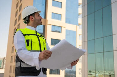 مفهوم وادرة المشاريع وتدقيقها، الأشراف على العمل لمتابعة سير المشاريع وتنفيذها، صورة جانبية لمهندس عربي خليجي سعودي يرتدي الثوب التقليدي والسترة الخاصة وخوذة الحماية يحمل مخطط هندسي ويتأمل التفاصيل الهندسية للبرج، خلفية أبراج وناطحات سحاب