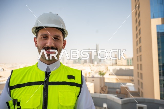 العمل في المجال الهندسي، الوقوف بفخر واعتزاز والنظر إلى الكاميرا, صورة مقربة لمهندس عربي خليجي سعودي مبتسم يرتدي سترة العمل وخوذة الحماية البيضاء, ، الاهتمام بالتطور العمراني بسواعد المهندسين السعوديين، خلفية الأبراج وناطحات السحاب في مدينة الرياض