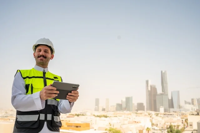   تقدم وتطور الهندسة المعمارية على ايدي المبدعين أبناء الوطن، نمو القطاع الهندسي في المملكة، مهندس عربي خليجي سعودي يرتدي الثوب التقليدي والسترة الخاصة وخوذة الحماية يقف ويمسك الهاتف المحمول يتابع العمل حسب استراتيجية سير المشروع، خلفية أبراج وناطحات سحاب