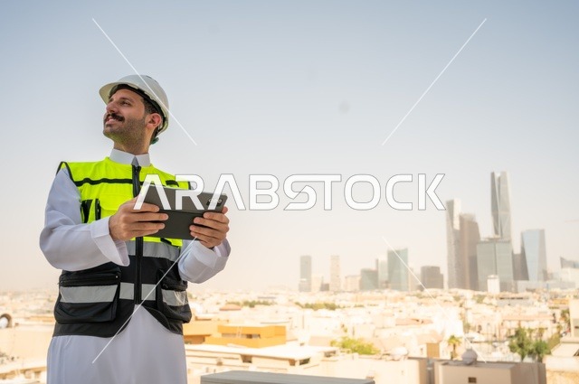   الإعمار بسواعد أبناء الوطن، تطور ونمو القطاع الهندسي في المملكة، متابعة العمل حسب استراتيجية سير المشروع، صورة جانبية لمهندس عربي خليجي سعودي يرتدي الثوب التقليدي والسترة الخاصة وخوذة الحماية يقف ويمسك الهاتف المحمول، خلفية أبراج وناطحات سحاب