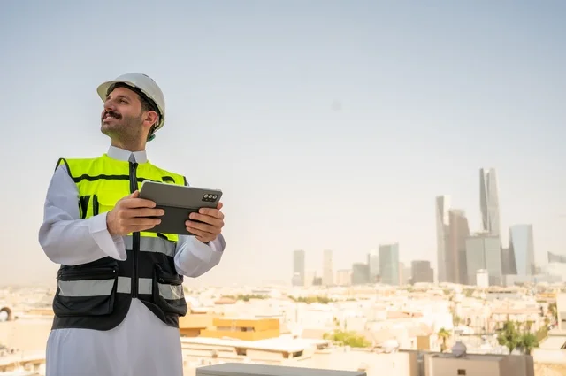   الإعمار بسواعد أبناء الوطن، تطور ونمو القطاع الهندسي في المملكة، متابعة العمل حسب استراتيجية سير المشروع، صورة جانبية لمهندس عربي خليجي سعودي يرتدي الثوب التقليدي والسترة الخاصة وخوذة الحماية يقف ويمسك الهاتف المحمول، خلفية أبراج وناطحات سحاب