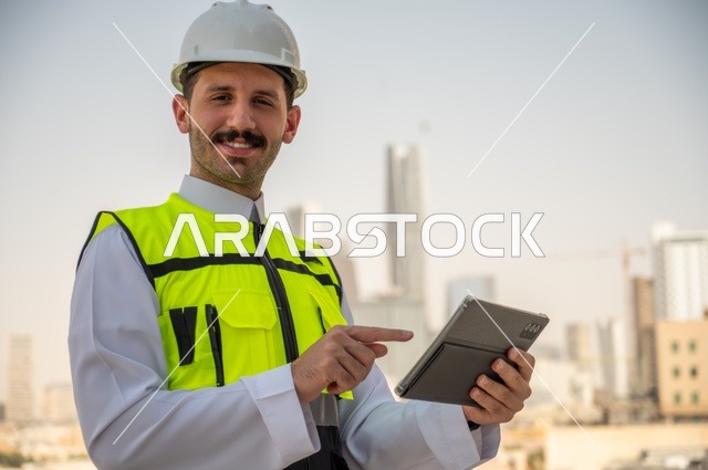   الإعمار بسواعد أبناء الوطن، تطور ونمو القطاع الهندسي في المملكة، متابعة العمل حسب استراتيجية سير المشروع، صورة جانبية لمهندس عربي خليجي سعودي يرتدي الثوب التقليدي والسترة الخاصة وخوذة الحماية يقف ويمسك الهاتف المحمول، خلفية أبراج وناطحات سحاب