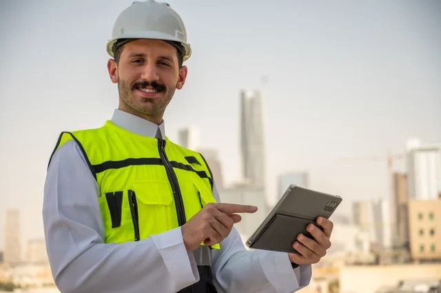 Reconstruction by the hands of the sons of the nation, development and growth of the engineering sector in the Kingdom, follow-up of work according to the project progress strategy, a side view of a Saudi Arabian Gulf engineer wearing traditional clothing, a special jacket and a protective helmet, standing and holding a mobile phone, a background of towers and skyscrapers