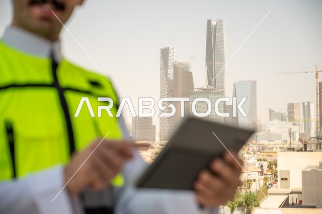  تقدم القطاع الهندسي في المملكة، نمو وتطور الهندسة المعمارية على ايدي المبدعين أبناء الوطن،صورة جانبية مقربة لمهندس عربي خليجي سعودي يرتدي الثوب التقليدي والسترة الخاصة وخوذة الحماية يقف ويحمل الهاتف المحمول، خلفية أبراج وناطحات سحاب