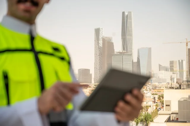 The progress of the engineering sector in the Kingdom, the growth and development of architecture at the hands of the creative sons of the nation, a close-up side view of a Saudi Arabian Gulf engineer wearing traditional clothing, a special jacket and a protective helmet, standing and holding a mobile phone, a background of towers and skyscrapers