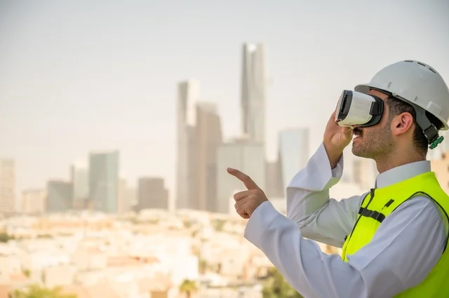  العمل في القطاع الهندسي، نمو وتطور الهندسة المعمارية على ايدي المبدعين أبناء الوطن، صورة جانبية لمهندس عربي خليجي سعودي يرتدي الثوب التقليدي والسترة الخاصة وخوذة الحماية ينظر في نظارة واقع افتراضي ثلاثية الأبعاد، خلفية أبراج وناطحات سحاب 