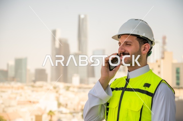 Background of towers and skyscrapers in Riyadh, the progress of the engineering sector in the Kingdom, the growth and development of architecture at the hands of creative sons of the nation, a side view of a Saudi Arabian Gulf engineer wearing traditional clothing, a special jacket and a protective helmet, standing and talking on a mobile phone
