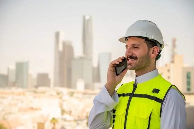 Background of towers and skyscrapers in Riyadh, the progress of the engineering sector in the Kingdom, the growth and development of architecture at the hands of creative sons of the nation, a side view of a Saudi Arabian Gulf engineer wearing traditional clothing, a special jacket and a protective helmet, standing and talking on a mobile phone