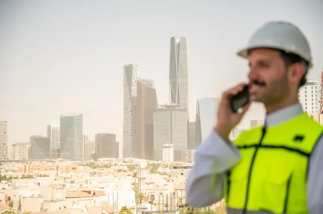  القطاع الهندسي المتطور في المملكة، نمو وتطور الهندسة المعمارية على ايدي المبدعين أبناء الوطن، صورة جانبية لمهندس عربي خليجي سعودي يرتدي الثوب التقليدي والسترة الخاصة وخوذة الحماية يقف ويتحدث من خلال الهاتف المحمول، خلفية أبراج وناطحات سحاب في الرياض