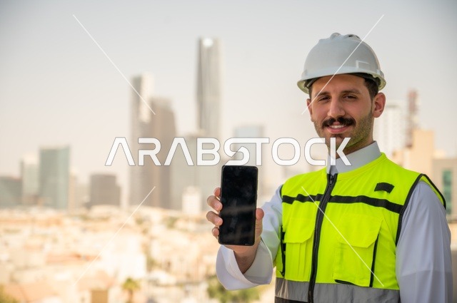 Advancement and development of architecture at the hands of creative sons of the nation, growth of the engineering sector in the Kingdom, a Saudi Arabian Gulf engineer wearing traditional clothing, a special jacket and a protective helmet stands and holds a mobile phone following up on work according to the project progress strategy, background of towers and skyscrapers