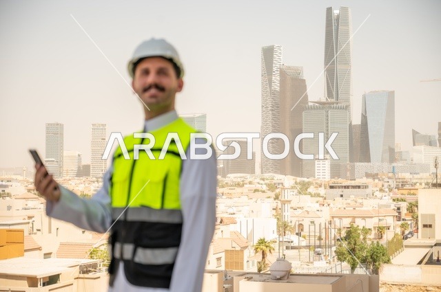 متابعة العمل حسب استراتيجية سير المشروع، تطور ونمو القطاع الهندسي في المملكة، الإعمار بسواعد أبناء الوطن، صورة جانبية لمهندس عربي خليجي سعودي يرتدي الثوب التقليدي والسترة الخاصة وخوذة الحماية يقف ويمسك الهاتف المحمول، خلفية أبراج وناطحات سحاب