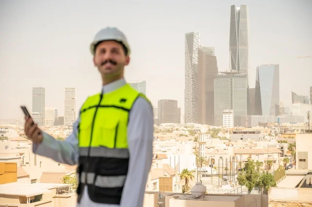 Follow-up of work according to the project progress strategy, development and growth of the engineering sector in the Kingdom, reconstruction by the hands of the sons of the homeland, a side view of a Saudi Arabian Gulf engineer wearing traditional clothing, a special jacket and a protective helmet, standing and holding a mobile phone, a background of towers and skyscrapers