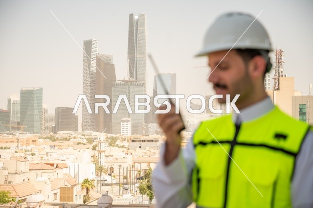 Following the progress of construction projects, the growth and development of architecture at the hands of creative sons of the nation, a side view of a Saudi Arabian Gulf engineer wearing traditional clothing, a special jacket, and a protective helmet, standing and talking on a cordless phone, with a background of towers and skyscrapers in Riyadh.