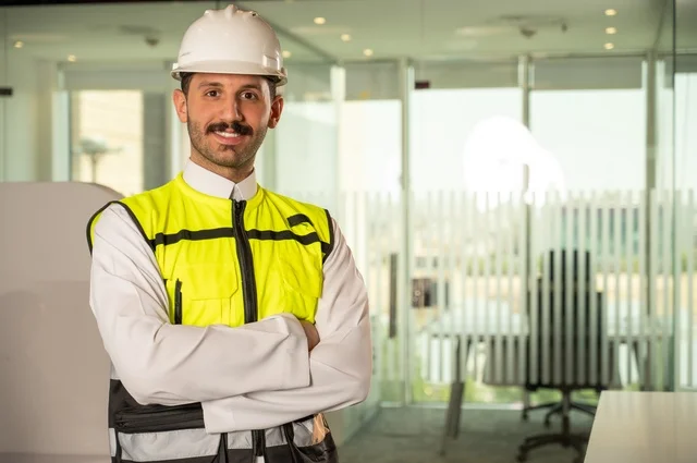  الوقوف بفخر واعتزاز والنظر إلى الكاميرا, التطور العمراني بسواعد المهندسين السعوديين، صورة مقربة لمهندس عربي خليجي سعودي مبتسم يرتدي سترة العمل وخوذة الحماية البيضاء,ايماءات أيدي متقاطعة, خلفية داخل مكتب هندسي