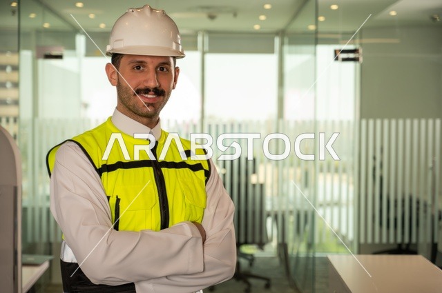  الوقوف بفخر واعتزاز والنظر إلى الكاميرا, التطور العمراني بسواعد المهندسين السعوديين، صورة مقربة لمهندس عربي خليجي سعودي مبتسم يرتدي سترة العمل وخوذة الحماية البيضاء,ايماءات أيدي متقاطعة, خلفية داخل مكتب هندسي