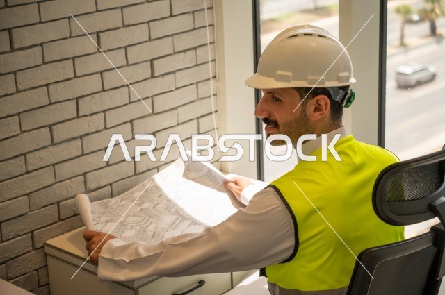 Close-up side view of a Saudi Arabian Gulf engineer wearing traditional thobe, jacket and work helmet sitting at a desk holding an engineering plan, following up and checking the progress of the project, engineering and construction concept, completing the construction drawings