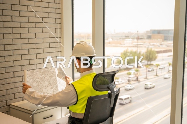 متابعه وتدقيق سير المشروع, مفهوم الهندسة والإنشاءات، صورة خلفية مقربة لمهندس عربي خليجي سعودي يرتدي الثوب التقليدي والسترة والخوذة الخاصة بالعمل يجلس على المكتب ويمسك بيده المخطط الهندسي ، إكمال رسم مخطوطات البناء