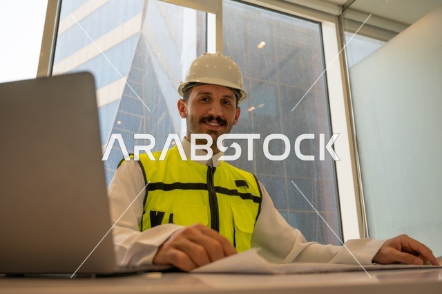 الإمساك بمخططات الإعمار، صورة مقربة لمهندس عربي خليجي سعودي مبتسم يرتدي الثوب التقليدي والسترة والخوذة الخاصة بالعمل يجلس على المكتب ويمسك بيده المخطط الهندسي بجانبه الحاسب المحمول، متابعه وتدقيق سير المشروع, مفهوم الهندسة والإنشاءات