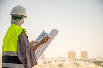 Reviewing and checking engineering plans, side view of a Saudi Arabian Gulf female architect holding a building plan, a veiled female construction engineer wearing a safety suit at work, formal work clothes, work helmet, construction project, background of buildings and structures