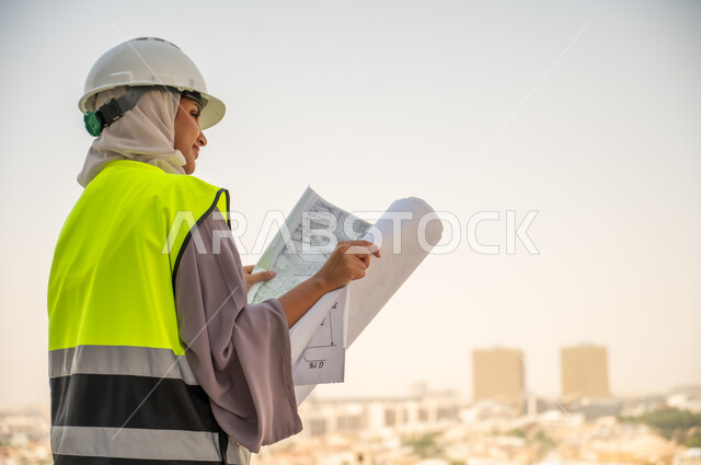  صورة جانبية لمهندسة معمارية عربية سعودية خليجية تحمل بيدها مخطط البناء ، مهندسة انشاءات محجبة ترتدي بدلة السلامة في العمل، لباس رسمي للعمل ، خوذة العمل,مراجعة المخططات الهندسية وتدقيقها, مشروع بناء ، خلفية عمارات وأبنية