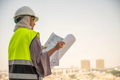 Side view of a Saudi Arabian Gulf female architect holding a building plan, a veiled female construction engineer wearing a safety suit, formal work clothes, work helmet, reviewing and checking engineering plans, construction project, background of buildings and structures