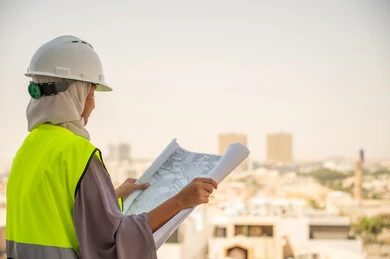 مراجعة المخططات الهندسية وتدقيقها, صورة جانبية لمهندسة معمارية عربية سعودية خليجية تحمل بيدها مخطط البناء ، مهندسة انشاءات محجبة ترتدي بدلة السلامة في العمل، لباس رسمي للعمل ، خوذة العمل, مشروع بناء ، خلفية عمارات وأبنية