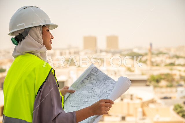  مشروع بناء ، خلفية عمارات وأبنية، صورة جانبية لمهندسة معمارية عربية سعودية خليجية تحمل بيدها مخطط البناء وتتأمل معالم المدينة ، مهندسة انشاءات محجبة ترتدي بدلة السلامة في العمل، لباس رسمي للعمل ، خوذة العمل, مراجعة المخططات الهندسية وتدقيقها
